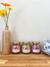 Three jars of tea with floral labels on a wooden surface next to a vase with flowers.